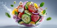 Fresh vegetable salad with cucumber, tomato, radish and herbs splashing in glass bowl on gray background with flying ingredients and water droplets.