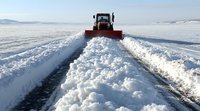 Snow plow clearing winter road during heavy snowfall, creating path through deep snow with high banks on sides against cloudy sky and mountain landscape.