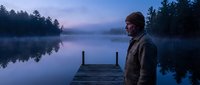 A middle-aged Caucasian male wearing a brown beanie and jacket stands on a wooden dock at dawn, gazing across a misty lake surrounded by evergreen trees and calm reflective waters.