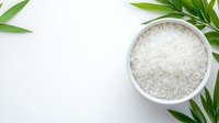 Raw white jasmine rice in ceramic bowl with green bamboo leaves on white background, overhead view for food photography and menu design.