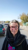 Senior woman wearing pink-framed glasses and black headscarf smiles at camera in outdoor parking lot setting with clear blue sky and blurred vehicles background.