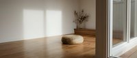 Minimalist modern interior with natural light streaming through large windows onto warm wooden floors. Woven basket and dried branches create serene atmosphere.