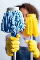 Person in yellow rubber gloves holding blue and yellow mops for cleaning, focus on cleaning tools against white background.