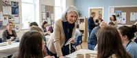Senior female teacher with gray hair in beige blazer helping diverse students in modern classroom with bulletin boards and natural lighting.