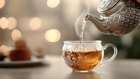Steaming hot tea poured from ornate silver teapot into transparent glass cup with delicate floral pattern against soft bokeh background, creating cozy atmosphere.