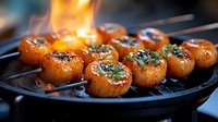 Grilled scallops with herbs on black cast iron pan, flames in background creating caramelized glaze, garnished with fresh chopped parsley and seasonings.
