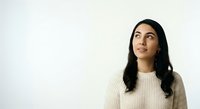 Young woman with long black hair and South Asian ethnicity wearing a cream-colored knit sweater, looking upward thoughtfully against a white background. Suitable for lifestyle, wellness, and inspirational content.