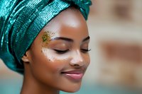 Young African woman wearing emerald green turban headwrap with glitter makeup and gold leaf decoration on face, eyes closed in serene expression against blurred background.