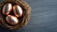 Three copper metallic eggs with decorative patterns nestled in rustic straw nest against dark gray wooden background, dramatic side lighting creates depth.