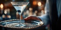 Elegant sparkling cocktail in martini glass served on silver tray by waiter in upscale bar with atmospheric bokeh background.