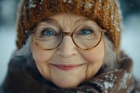 Senior woman with fair skin, Caucasian ethnicity, wearing round eyeglasses and a brown knit beanie, smiling warmly in snowy winter conditions. Suitable for healthcare, lifestyle, and seasonal campaigns.