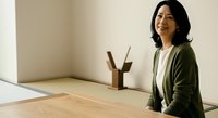 Asian woman in olive cardigan standing at wooden desk with pencil holder in minimalist office space, suitable for business, corporate, and professional lifestyle content.