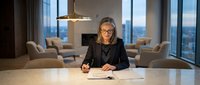 Professional businesswoman with glasses reviewing documents at conference table in modern office with city view during evening hours.