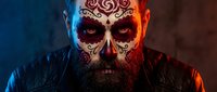Bearded man with intricate skull face paint featuring ornate swirl patterns in dramatic blue and orange lighting for Halloween or Day of the Dead themes.
