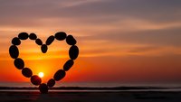 Heart-shaped arrangement of smooth stones silhouetted against vibrant sunset over ocean horizon. Romantic beach scene for love and wellness concepts.