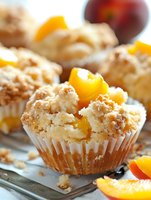 Fresh baked peach crumb muffins with streusel topping and juicy fruit pieces in white paper liners, extreme close up macro food photography on light surface.