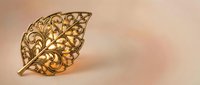 Ornate golden leaf brooch with intricate filigree scrollwork pattern on warm beige background for jewelry design and luxury fashion accessories.