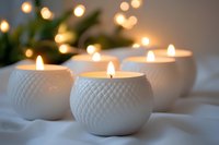 White ceramic candle holders with glowing tea lights creating cozy atmosphere on soft fabric. Decorative textured spherical vessels with warm bokeh lights.