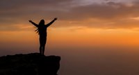 Woman silhouette with raised arms celebrating freedom on mountain cliff edge against dramatic orange sunset sky for motivational and success concepts.
