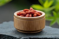 Goji berries in wooden bowl on slate surface with green plants background for healthy superfood nutrition and wellness lifestyle concepts.