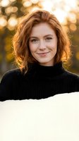Young redhead woman in black turtleneck sweater smiling warmly at camera with golden bokeh background during golden hour outdoor portrait session.