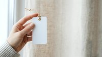 Hand holding blank white price tag or label with twine against neutral linen fabric background for product branding and retail display purposes.