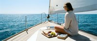 Woman with long brown hair, Caucasian ethnicity, wearing casual beige clothing, enjoying a meal on the bow of a sailboat during daytime on calm blue ocean waters.