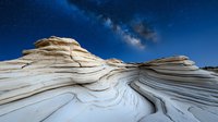 White sandstone rock formations with curved striped patterns under starry night sky and Milky Way galaxy, dramatic natural landscape in desert wilderness.