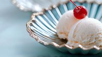 Vanilla ice cream scoop with maraschino cherry on top served in elegant silver dessert dish against soft blue background.