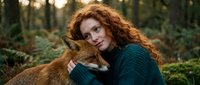 Young redhead woman with curly hair embracing red fox in forest setting. Cozy teal sweater, warm natural lighting, wildlife connection concept for nature campaigns.