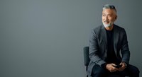 Mature male professional with gray hair and beard, wearing dark blazer and black shirt, seated against neutral gray background for corporate portraits and business headshots.