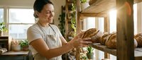 Mature male baker with dark hair and olive complexion holding fresh artisan bread loaves in rustic kitchen with natural sunlight, plants, and wooden beams for bakery, food business, and culinary content.