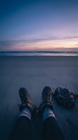 Traveler in hiking boots and backpack sitting on sandy beach at twilight, gazing at ocean horizon during golden hour sunset, adventure and wanderlust concept for travel.
