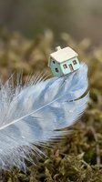Miniature house model balanced on delicate white feather among moss, symbolizing fragile dreams, home ownership aspirations, and lightweight living concepts.