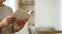 Senior woman with brown skin reading open book indoors, wearing beige sweater and gold ring, suitable for education, literacy, and lifestyle content.