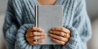 Woman in cozy blue knit sweater holding closed silver hardcover book, showcasing gold rings on her fingers.