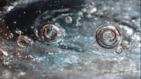 Macro photography of water droplets forming concentric circular ripples on dark blue-gray surface with light reflections creating abstract patterns.