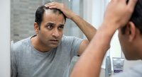 Middle-aged South Asian man examining hairline in bathroom mirror, touching forehead with concerned expression. Hair loss, balding, grooming concept.