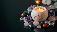 Lit candle in eggshell surrounded by colorful gemstones and crystals creating mystical spiritual atmosphere for meditation and healing practices.