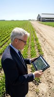 Senior Caucasian businessman using tablet computer for precision agriculture monitoring in green crop field with modern farm building background.