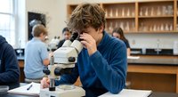 Young male student examining specimen through microscope in science laboratory classroom during biology lesson.