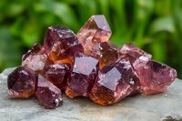 Raw purple fluorite crystal cluster with translucent cubic formations on stone surface against green natural background, showing mineral's natural geometric structure.