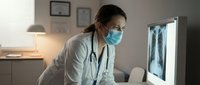 Female doctor in white coat and mask examines chest X-ray on monitor in clinic office, focused medical diagnosis concept.