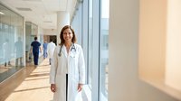 Confident young Hispanic female doctor in white coat with stethoscope standing in modern hospital corridor, smiling at camera.
