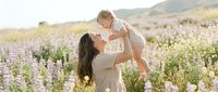 Young Caucasian mother lifting smiling baby in blooming lupine field at golden hour, joyful family moment outdoors in spring meadow.