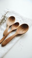 Three handcrafted wooden spoons arranged on white linen fabric with dried herbs, showcasing natural kitchen utensils for cooking and serving.