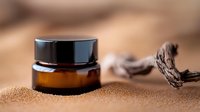 Small amber glass cosmetic jar with black lid on textured sand background with decorative driftwood piece, showcasing natural skincare packaging and spa elements.
