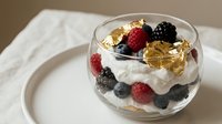 Layered berry parfait with fresh raspberries, blueberries, blackberries, whipped cream and golden granola in glass bowl on neutral background.