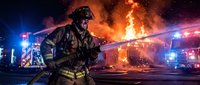 Firefighter in protective gear battles intense blaze at night scene with emergency vehicles and dramatic flames creating powerful rescue operation atmosphere.