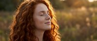 Young redhead woman with curly hair and freckles enjoying peaceful moment with eyes closed in warm golden sunlight outdoors during sunset or sunrise hour.